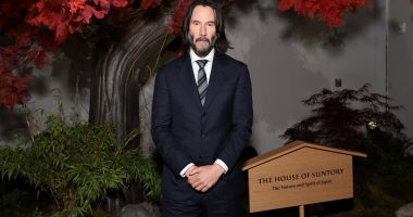 Keanu Reeves poses at an event with red tree leaves over his head and some shrubbery behind him